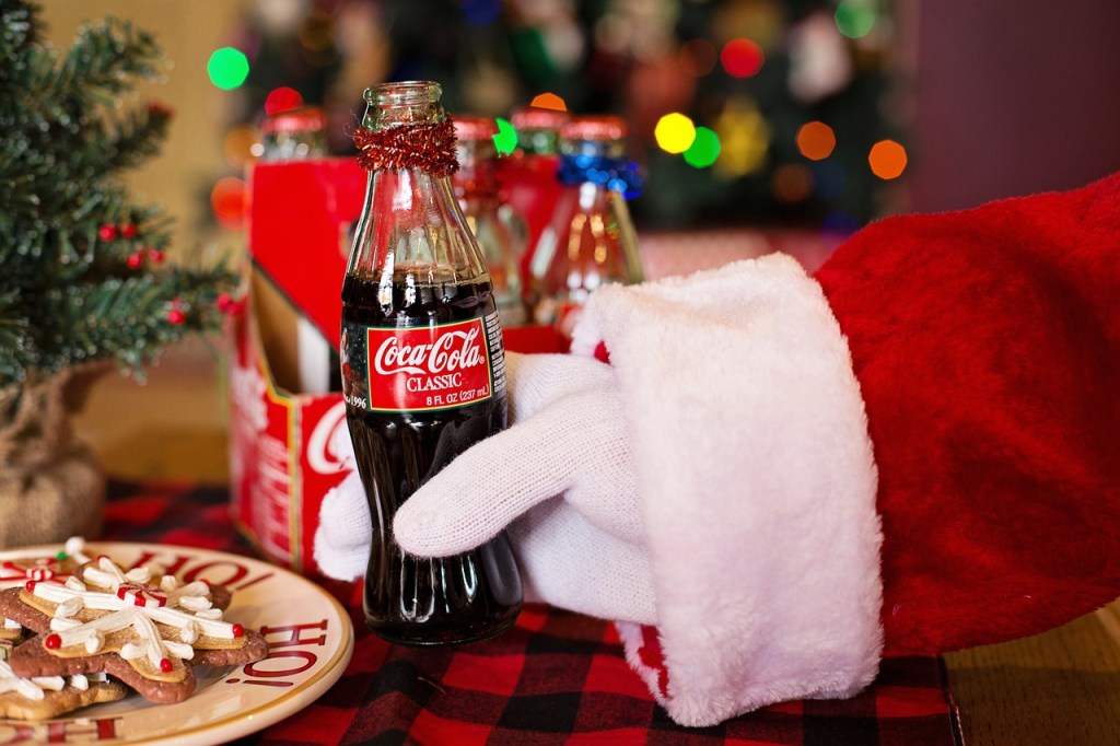 a white gloved hand with red and white sleeve picking up a bottle of coca cola, with Christmas decorations in the background