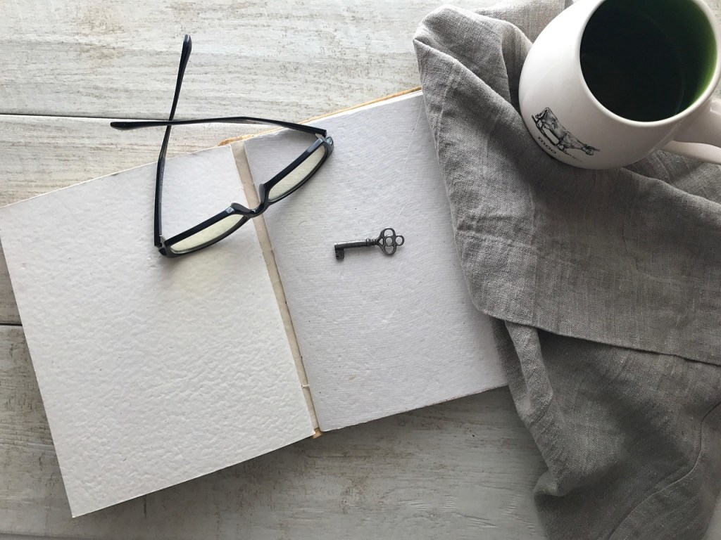 A white notebook with glasses and an old fashioned ornate key on top, next to some fabric with a white mug of coffee on it